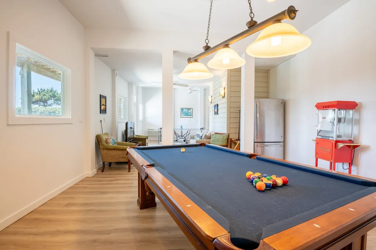 A vibrant game room featuring a pool table as the centerpiece, surrounded by bar-style light fixtures, comfortable seating, and a popcorn machine. The setup creates an inviting atmosphere for friendly competition and easy access to snacks.