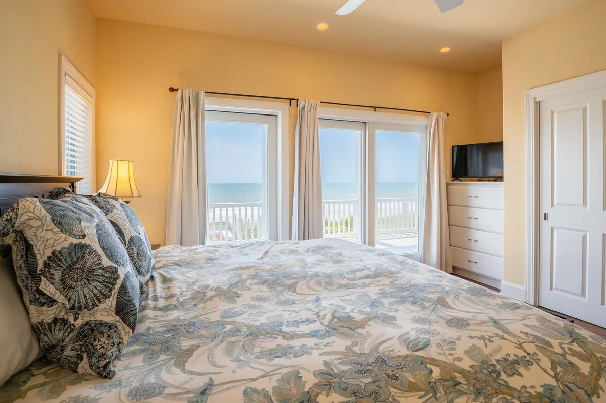A serene bedroom interior with a bed featuring floral bedding, large windows offering an ocean view, a dresser, and a TV. The room is filled with light and feels restful and inviting, underscoring the details about linens provided and beds freshly made.
