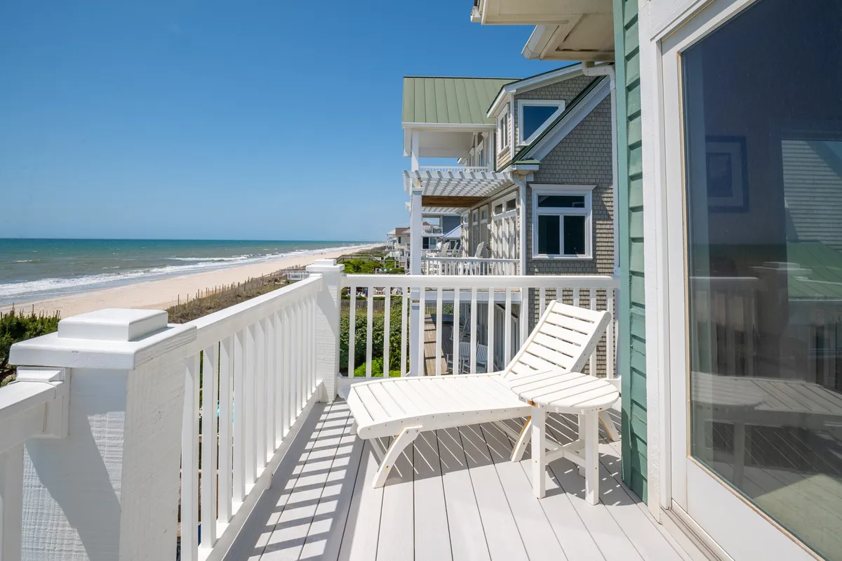 A tranquil balcony scene overlooking the beach, furnished with a lounge chair and bordered by a simple railing. The view emphasizes the property's proximity to the sand and surf, with opportunities for relaxation and beach exploration.