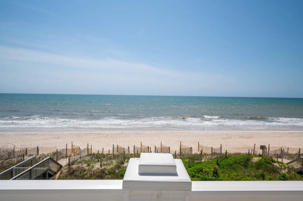 A picturesque view of ocean waves, sandy beach, and dune vegetation frames a boardwalk leading toward the water. The setting is peaceful and natural, evoking the exclusive, tranquil atmosphere of a private beachfront community.