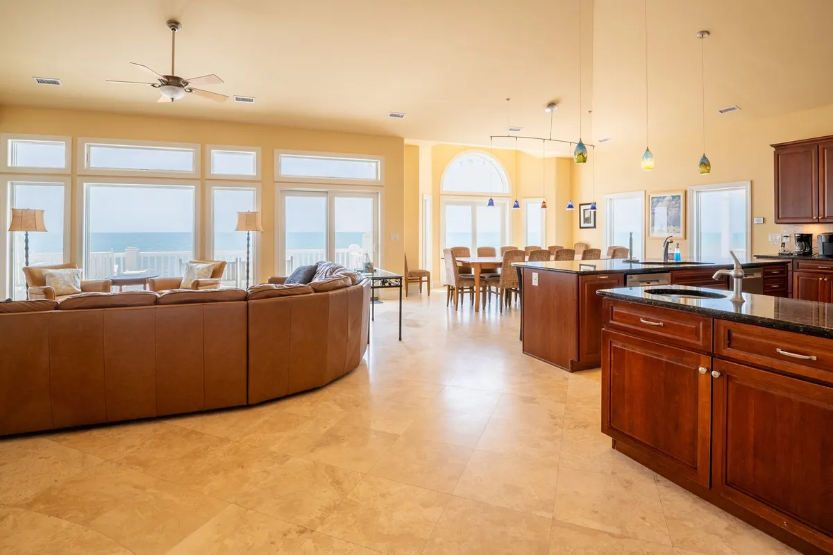 This image showcases an expansive, open-concept great room that seamlessly connects a large sectional sofa, dining table, and kitchen island. The space is bathed in natural light from oversized windows that offer sweeping ocean views, and the modern finishes reflect a sense of luxury and comfort. The visual connection between the various gathering spaces supports both intimate and group activities.