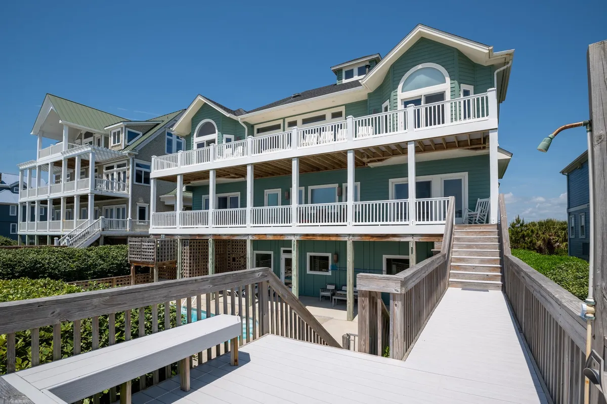 This exterior image features the multi-level deck of a custom home, showcasing the green exterior, large windows, and a staircase leading up. The scene is bathed in cheerful natural light, with hints of the lush landscaping surrounding the property. The atmosphere is relaxed, inviting, and hints at the exclusivity and tranquility of the neighborhood.