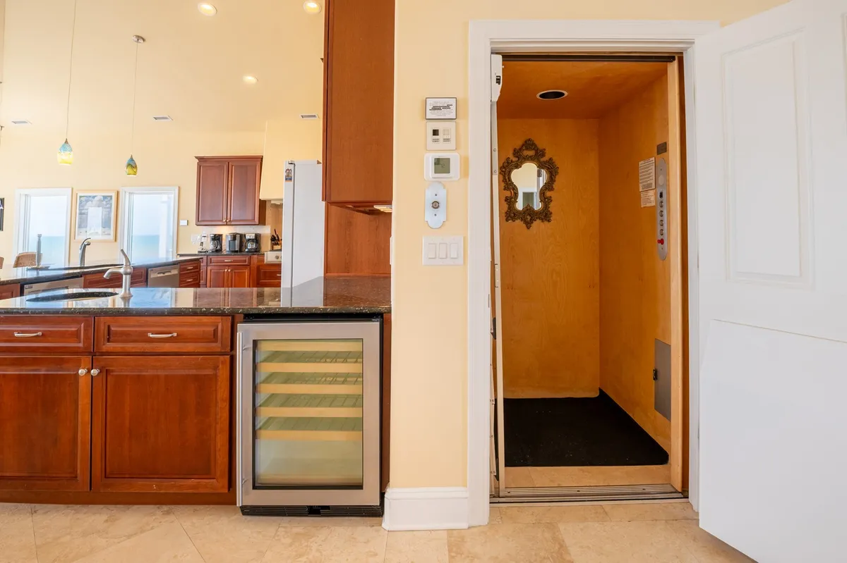 This image features a modern kitchen island with an elevator entrance in the background and a wine cooler. The setting exudes comfort, convenience, and upscale living, while the visible elevator directly addresses one of the section's highlighted amenities.