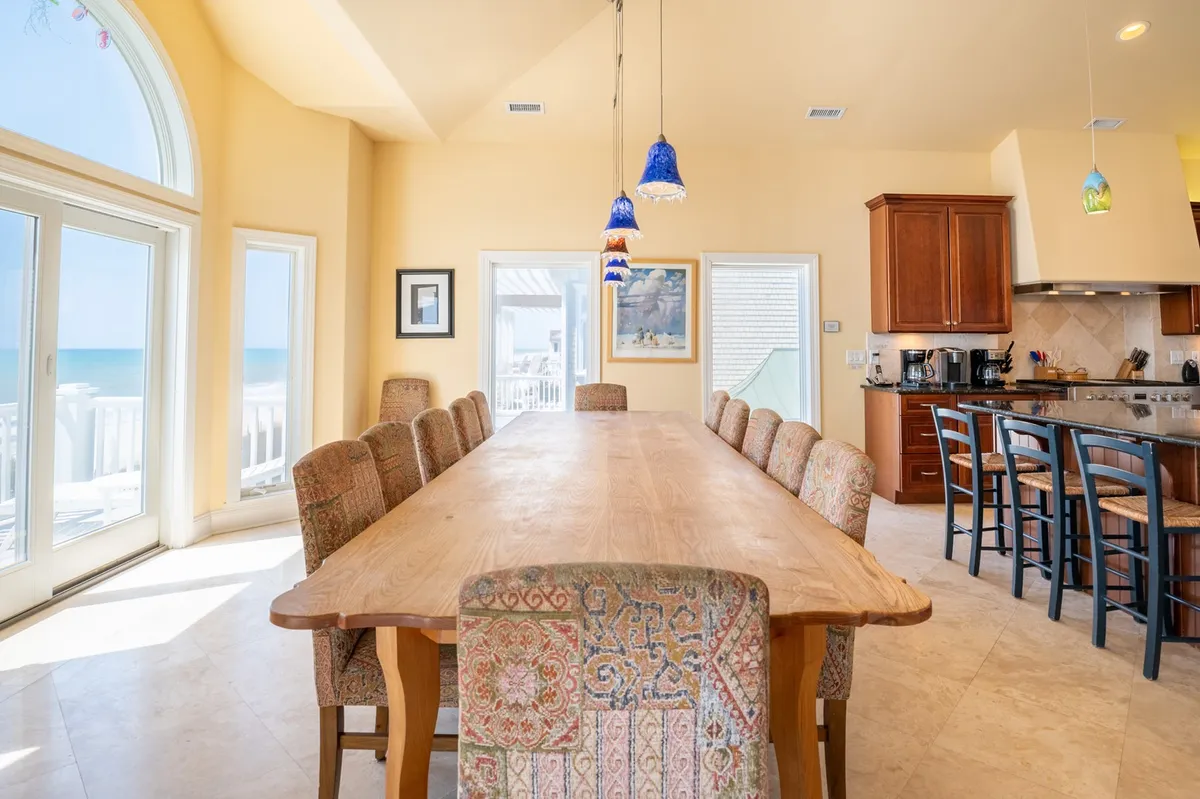 A striking view of a large, custom wooden dining table, surrounded by decorative chairs and colorful pendant lights. Panoramic windows provide a direct line of sight to the ocean, reinforcing the connection between indoor gatherings and outdoor beauty. The scene radiates warmth and is ideal for hosting large group dinners or special occasions.