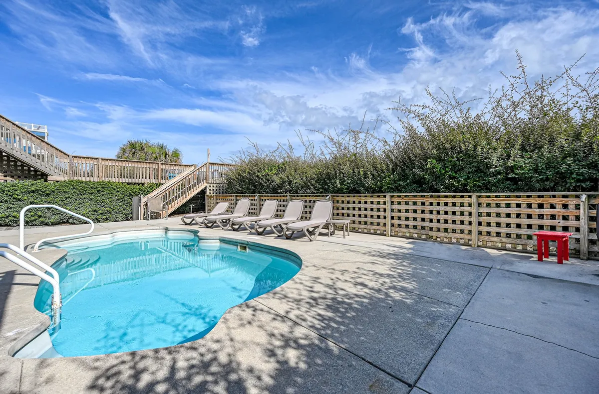 A sunlit poolside patio featuring a sparkling swimming pool, comfortable lounge chairs, and a vibrant red side table. The outdoor space encourages swimming, sunbathing, and relaxed social gatherings.