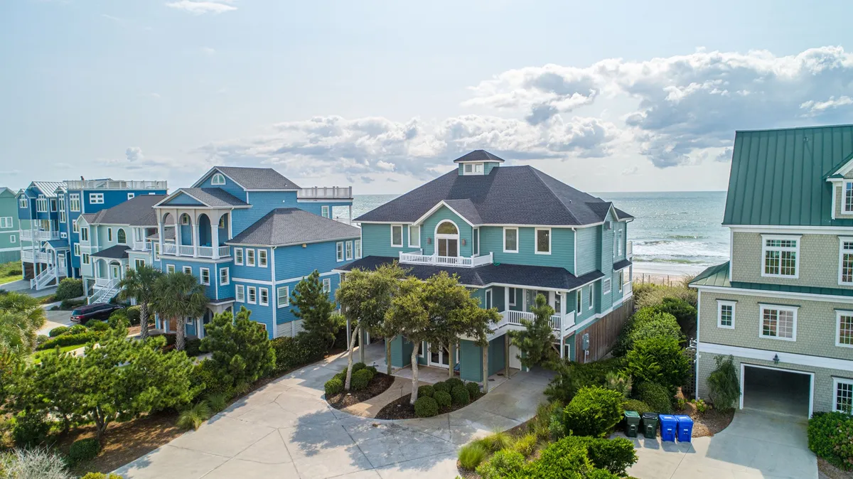 A spacious, inviting beachfront house set among palm trees with neighboring vibrant homes visible, evoking a relaxed coastal vibe. The porch and surrounding area hint at the proximity to the beach and easy outdoor access.