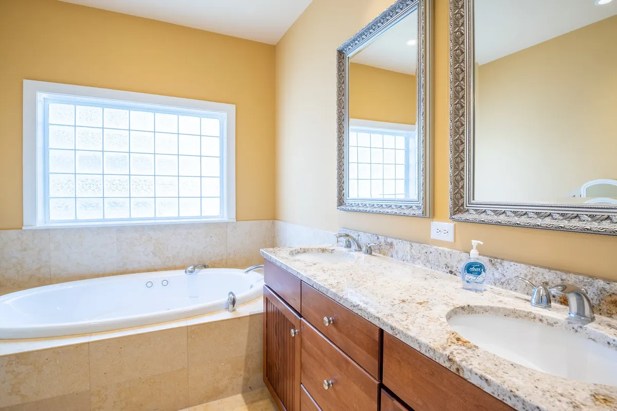 A serene bathroom with a bathtub, double sink vanity, large mirror, and window letting in soft natural light. The space exudes spa-like tranquility, ideal for both relaxing baths and morning routines.