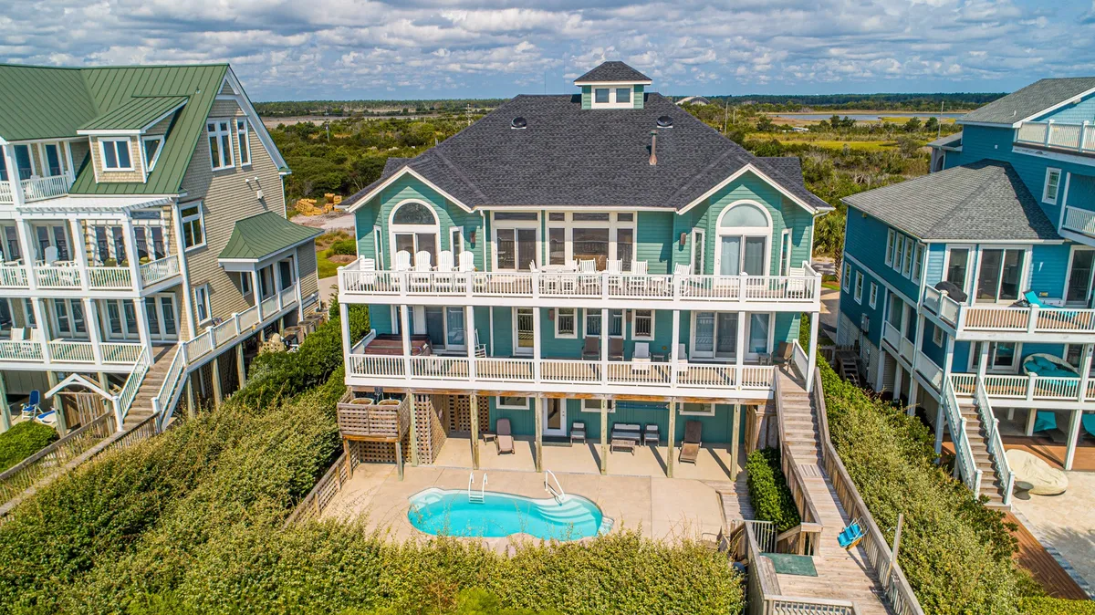 The image showcases the grand oceanfront estate's exterior, featuring a sparkling swimming pool surrounded by a spacious deck with lounge chairs, lush landscaping, and expansive windows overlooking the ocean. Sunset colors bathe the scene, highlighting inviting spaces for outdoor dining and relaxation, perfect for gathering with family and friends.