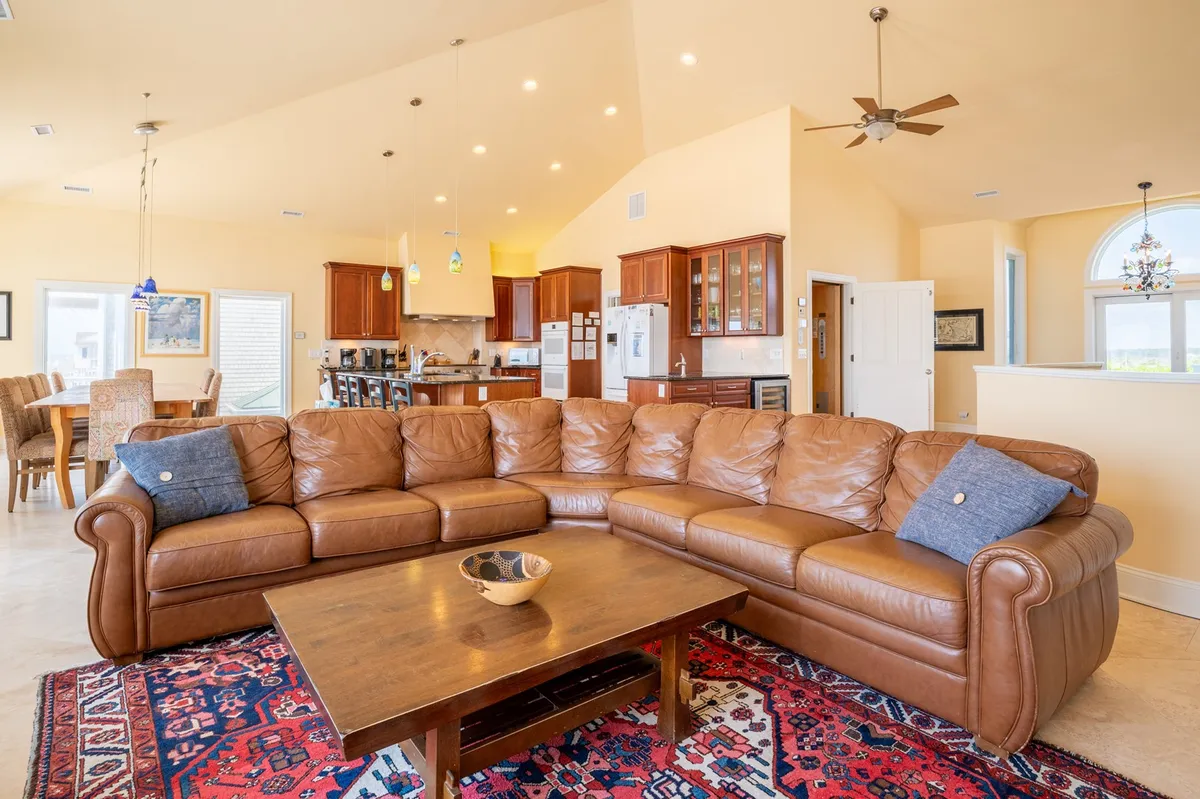 A cozy and vibrant scene featuring a leather sectional sofa, colorful area rug, and wood coffee table, with the kitchen area visible in the background. The design elements and layout underscore a comfortable, inviting space for socializing, relaxing, or celebrating with loved ones.