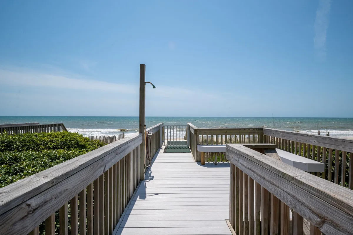 This image captures a wooden walkway leading directly to the beach, bordered by sand dunes and opening up to expansive ocean views under a blue sky. The scene evokes a sense of effortless access and the inviting tranquility of North Topsail Beach.