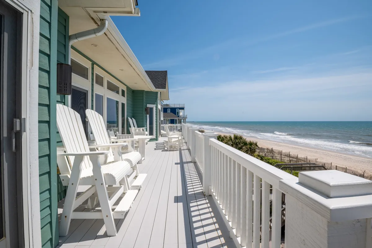 White Adirondack chairs line a porch overlooking the beach and the blue Atlantic, with the green exterior of a house anchoring the scene. The vibe is serene, and the view extends out to open ocean and sky, suggesting calm and exclusivity. The lush lawn and mature landscaping add to a sense of upscale comfort and tranquility.